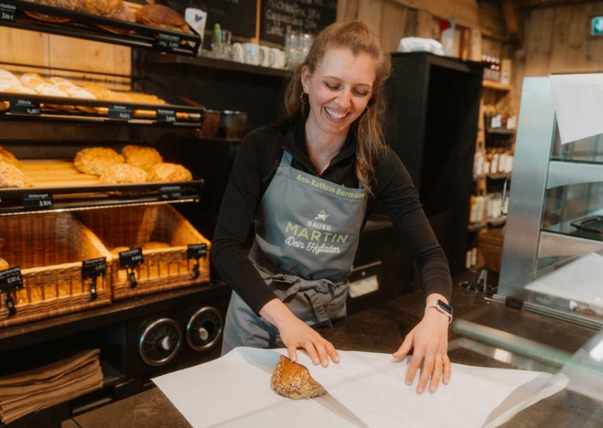 Verkäuferin mit Bauer-Martin-Schürze packt Brot ein, dahinter das Brotregal